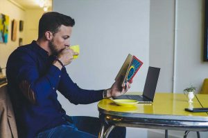 student sits on table while reading a book and drinking coffee